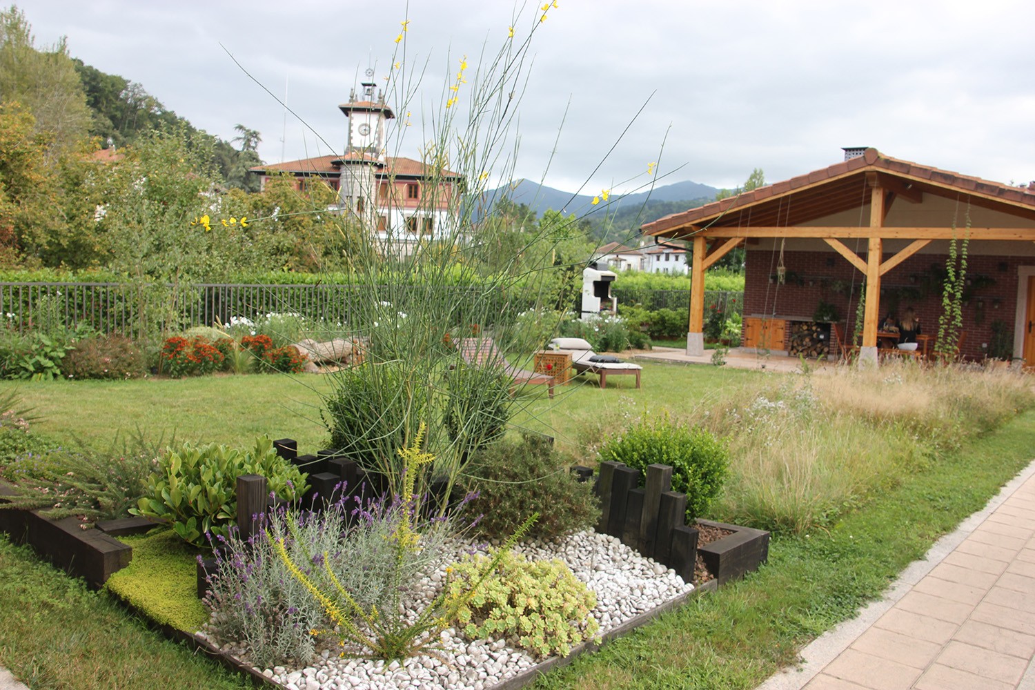 Jardín íntimo con parterre de grava blanca, plantas ornamentales, césped, pérgola y camino pavimentado.
