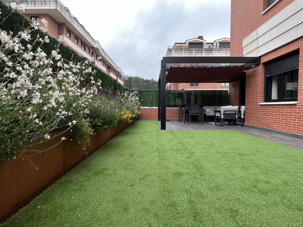 Rediseño de jardín con césped artificial, jardineras floridas y pérgola junto a terraza.