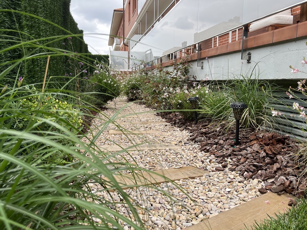 Sendero de losas y grava en jardín con plantas, balizas y seto junto a un edificio.