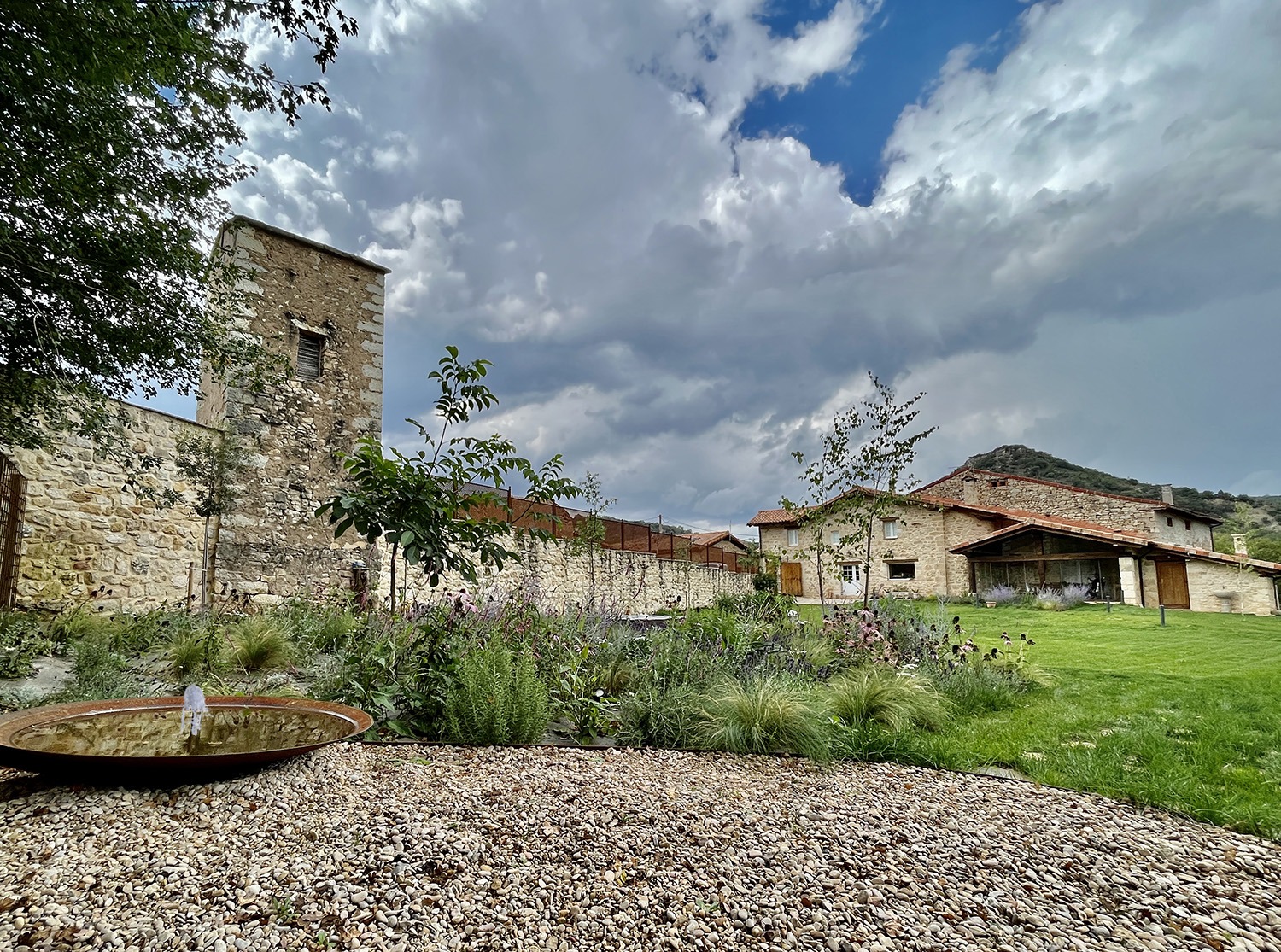 Organización de jardín con pradera, parterre de vivaces y fuente circular sobre grava junto a muro de piedra.
