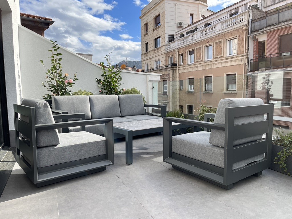 Terraza con sofá, sillones y mesa de exterior, jardineras y barandilla de cristal.