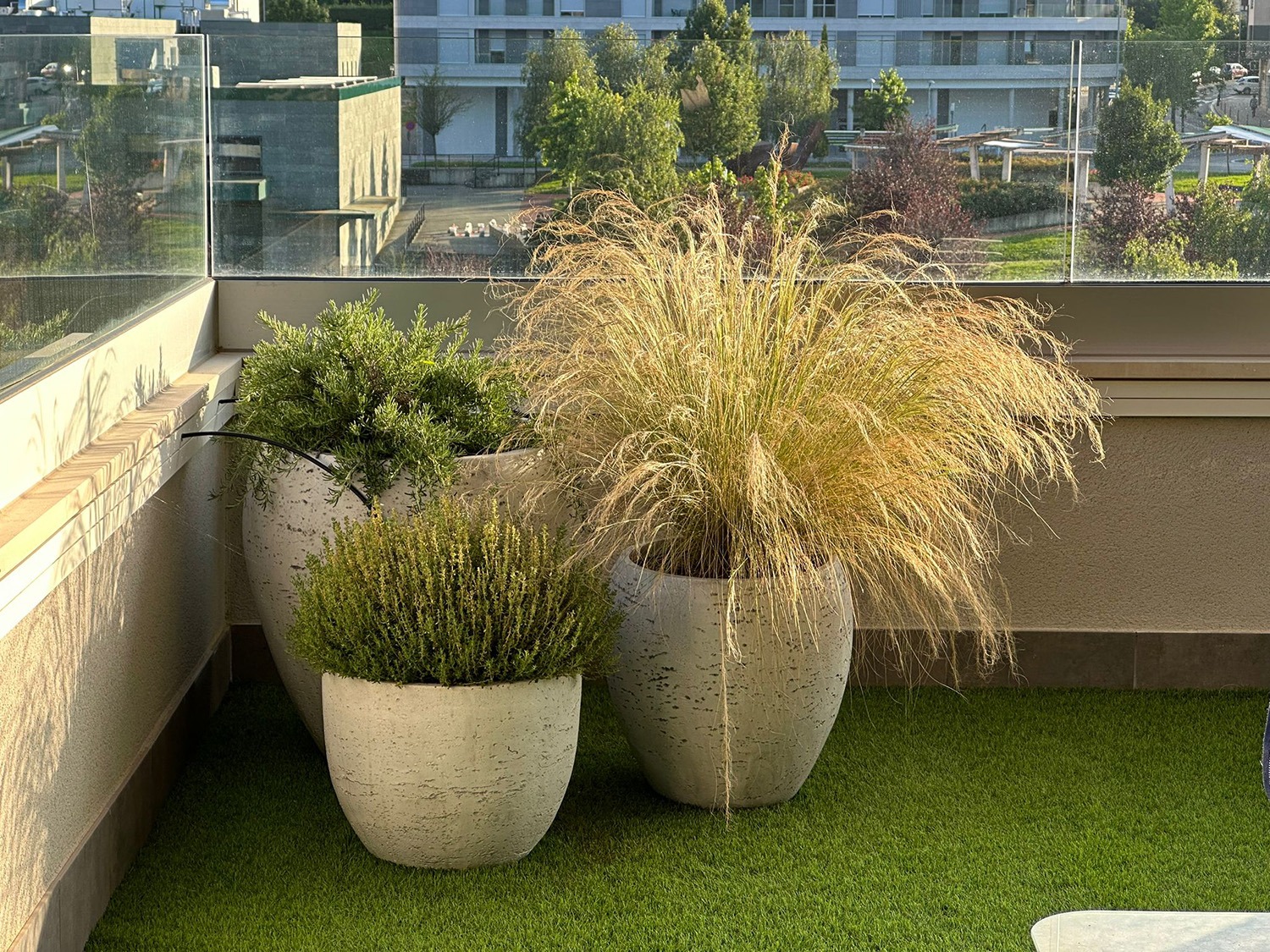 Terraza funcional con césped artificial, macetas claras con gramíneas y barandilla de cristal.