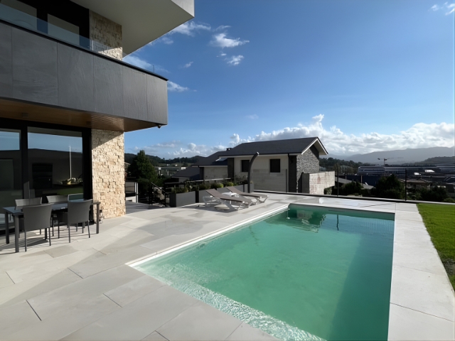 Jardín con piscina rectangular junto a vivienda moderna, terraza de piedra y tumbonas.