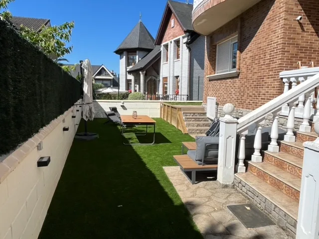 Jardín con césped artificial, mesa, sombrilla y escaleras junto a vivienda de ladrillo.
