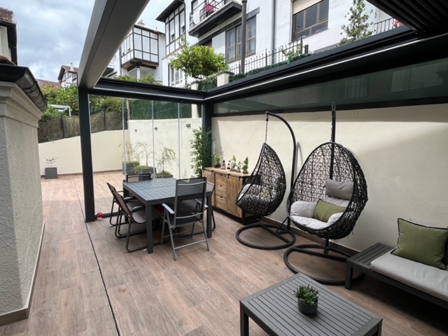 Terraza cubierta con tarima de madera, sofá, mesa y sillas, y dos sillas colgantes en el jardín.
