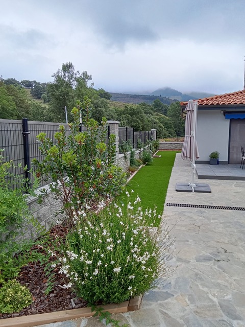 Jardín con terraza de piedra, parterre con flores blancas y césped junto a la casa.